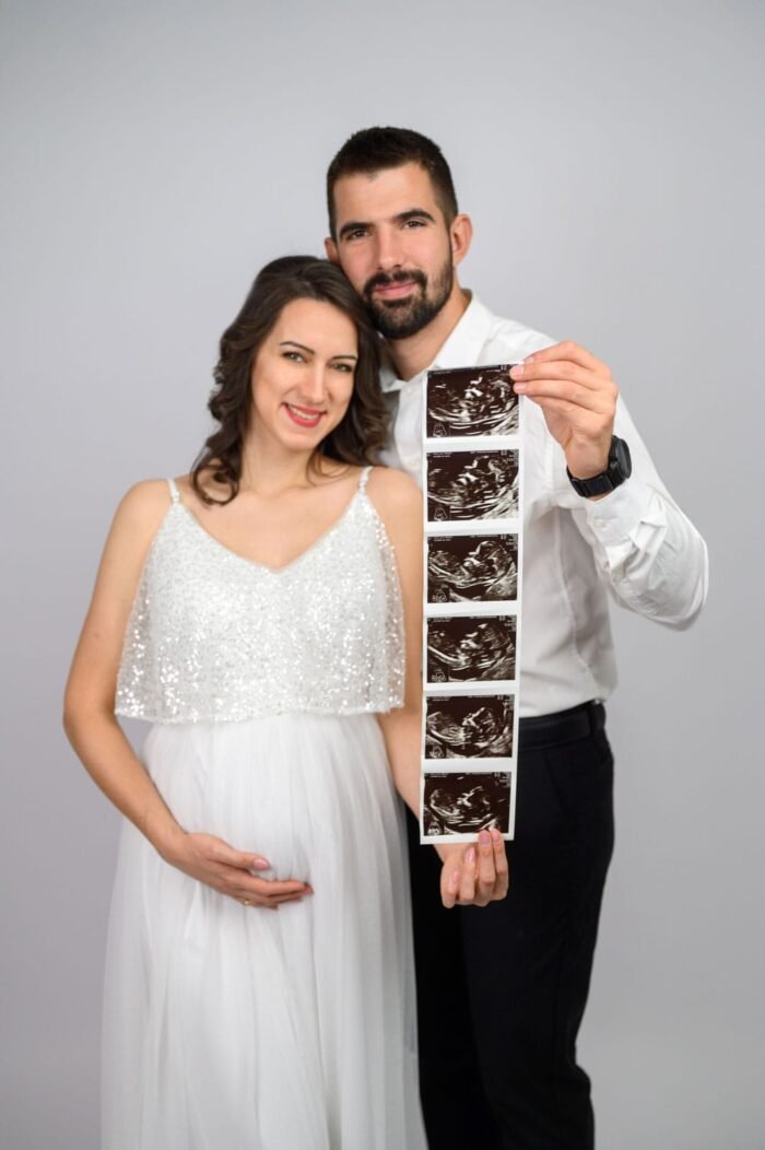 Várandós fotózás Debrecenben - Egy boldog pár mosolyogva tart egy ultrahang fotósorozatot a kamera felé, miközben a kismama kezét gyengéden a pocakjára helyezi. A kép a családalapítás örömét és a baba érkezésének izgatott várakozását tükrözi.