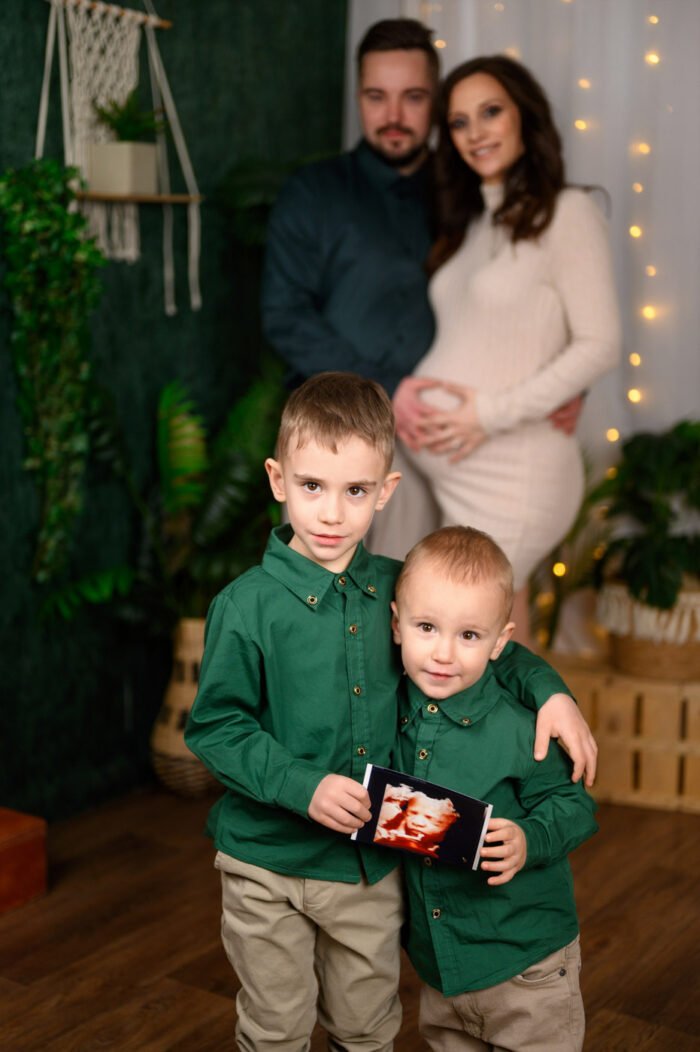 Családi kismama fotózás Debrecenben, ahol a nagyobb testvérek is osztoznak a babavárás örömében. A gyerekek kezükben tartják az ultrahangképet, miközben a szülők szeretetteljesen figyelik őket a háttérben. Stúdióban készült meghitt és érzelmes babaváró fotók.