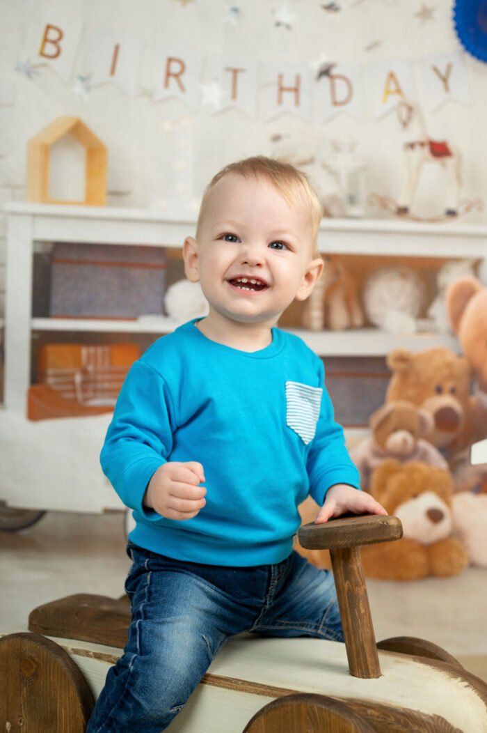 Vidám kisfiú nevet a kamerába, miközben egy vintage hintajátékon ül, ünnepi dekorációval a háttérben.