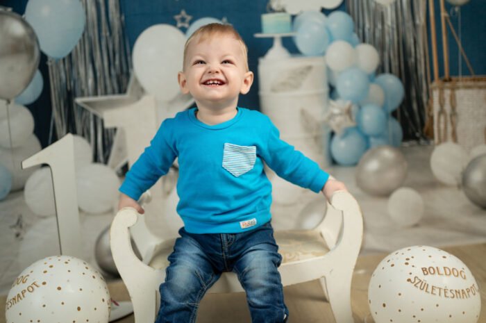 Kék pulóveres kisfiú ül egy fehér hintaszéken, körülötte lufik és csillagos szülinapi dekoráció.