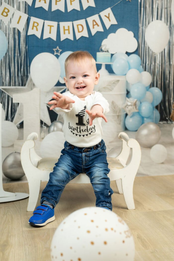 Kisfiú fehér bodyban boldogan nyúl a kamera felé, mögötte kék és fehér születésnapi dekoráció.