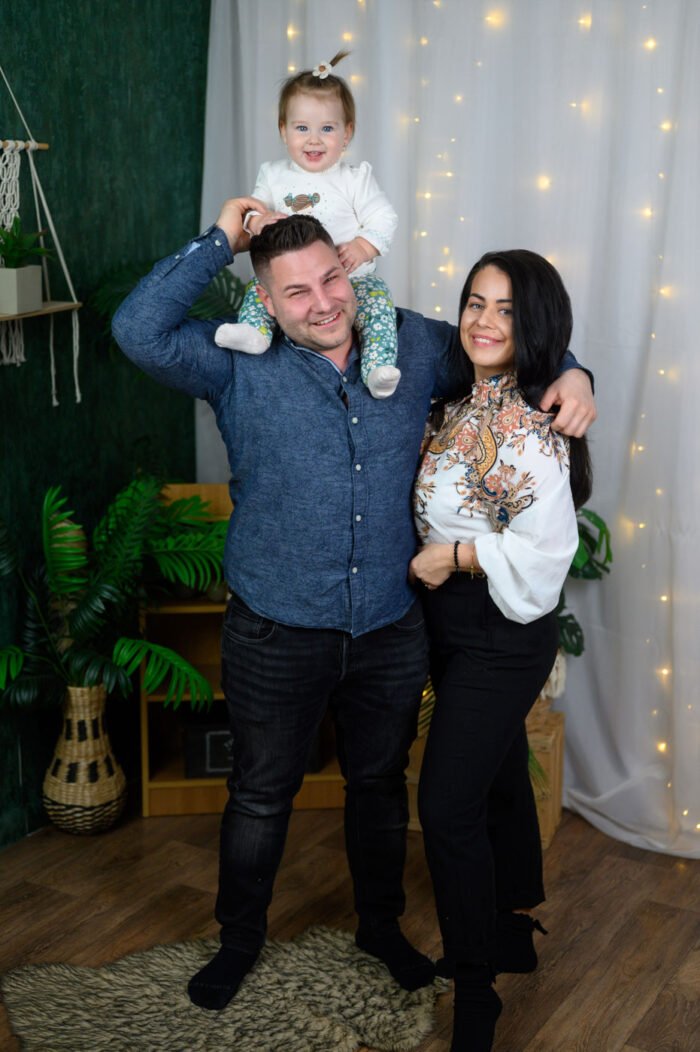Családi fotózás Debrecenben, ahol anya, apa és egyéves kislányuk együtt pózolnak meghitt, vidám hangulatban. Stúdiófotózás természetes dekorációval és fényfüzér háttérrel.