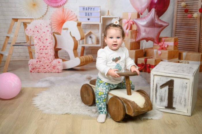 Egyéves kislány születésnapi fotózása Debrecenben, vintage játékautón ülve. Hangulatos dekoráció, rózsaszín lufik, ajándékdobozok és virágos szám egyes díszítés. Babafotózás stúdióban profi beállításokkal.