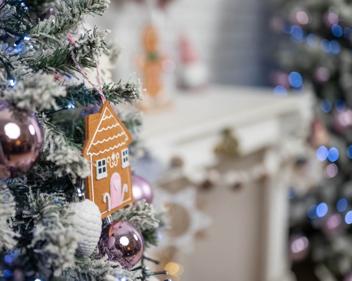 Közelkép egy mézeskalács házikó díszről, lilás karácsonyi gömbökkel és havas fenyőágakkal.