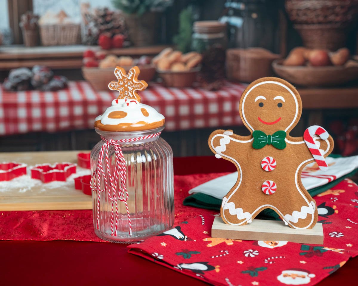 Mosolygós mézeskalács figura és hópelyhes díszüveg piros asztalon, karácsonyi háttérrel.