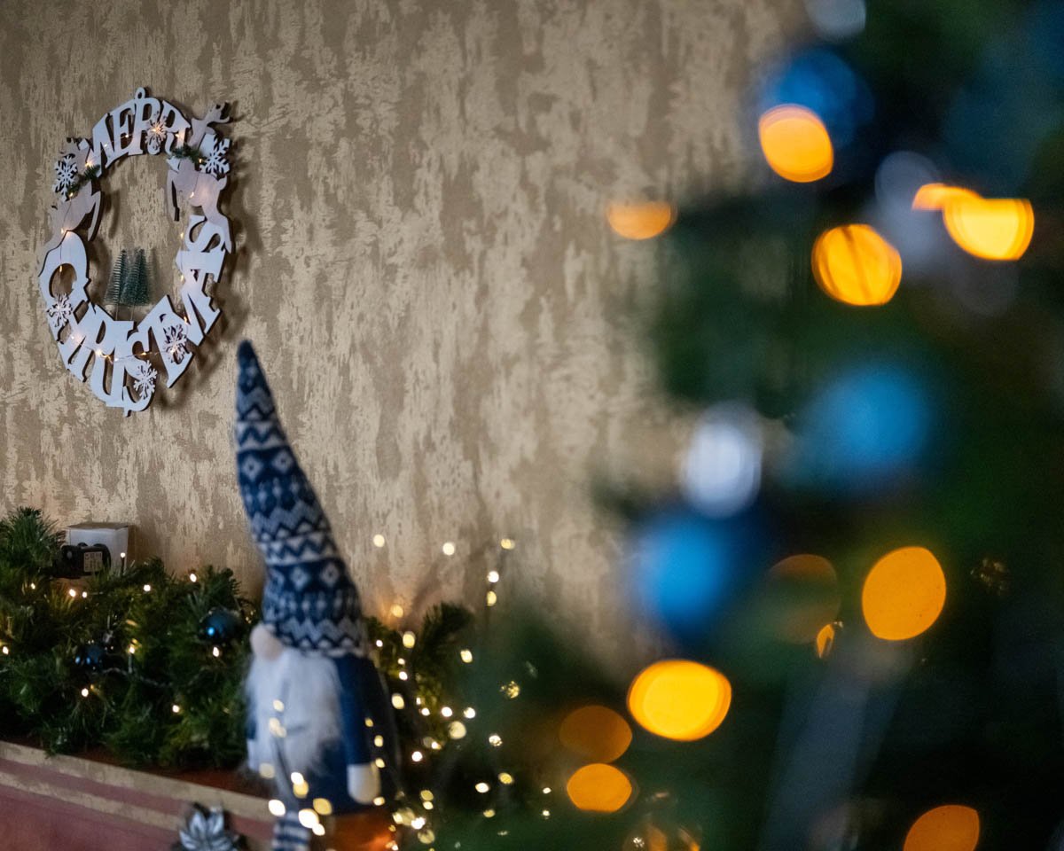 Fehér „Merry Christmas” koszorú arany falon, előtérben fényfüzérek és karácsonyi díszek.