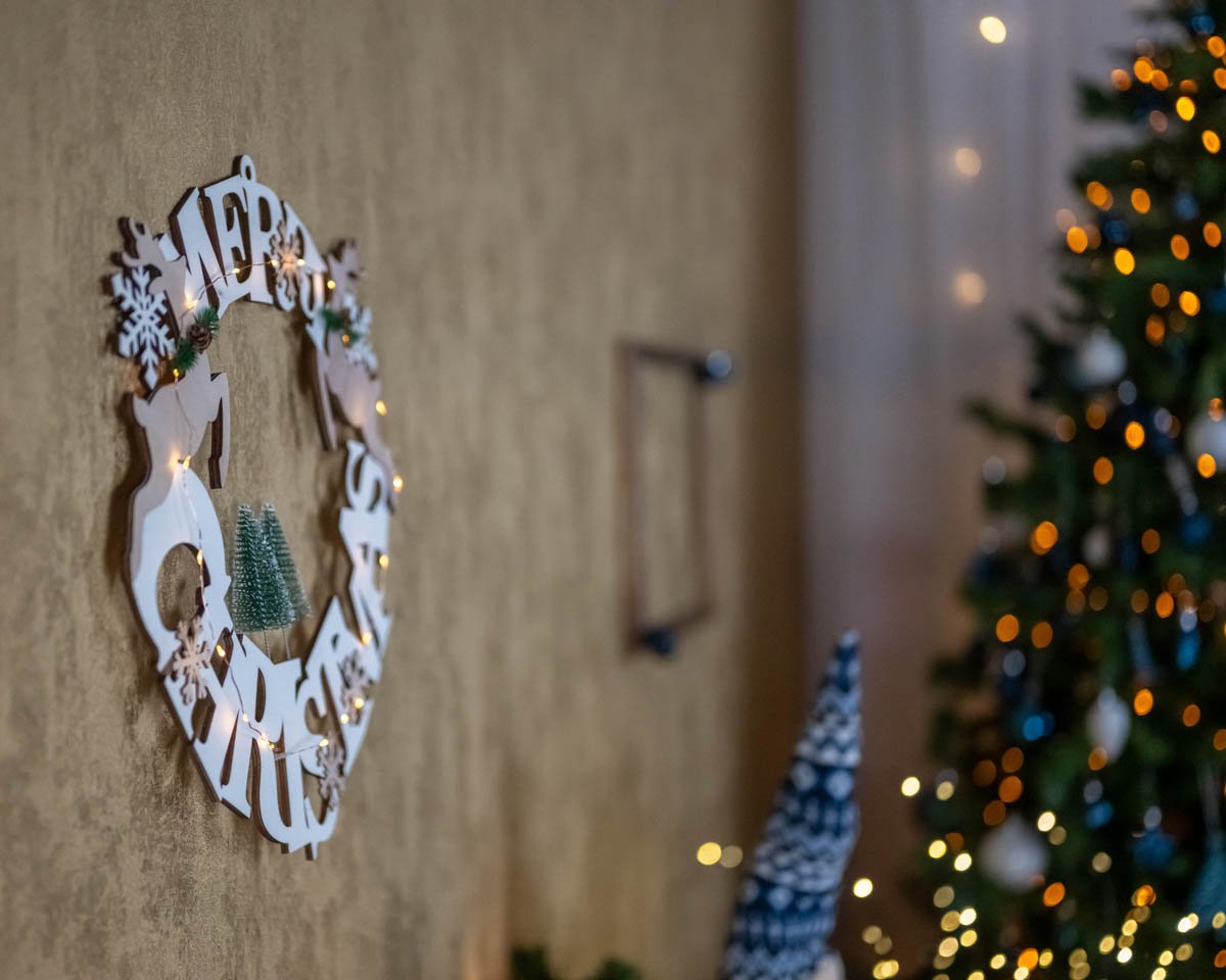 Fehér fa „Merry Christmas” koszorú a falon, háttérben karácsonyfa fényekkel.