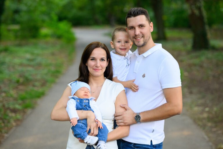Édesanya a kisbabát, édesapa a kisfiút tartja a karjában egy parki ösvényen, fehér-farmer összeállításban