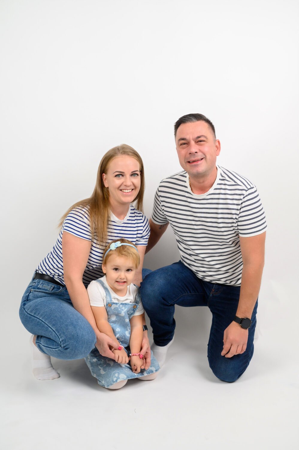 Anya, apa és kislányuk fehér hátterű stúdióban, csíkos pólóban és farmerben, mosolyogva.