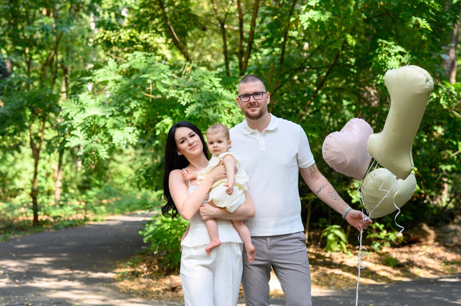 Háromtagú család a parkban: anya a kislányát tartja, apa szív és egyes alakú lufikkal, nyári szabadtéri fotózás.