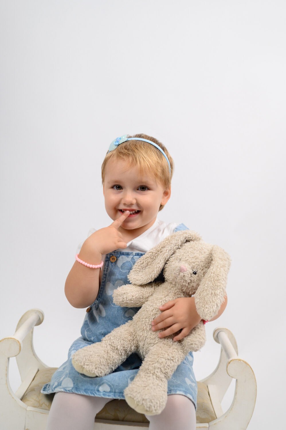 Kislány fehér padon ülve plüss nyuszit tart, ujját a szájához érintve, vidáman mosolyogva.