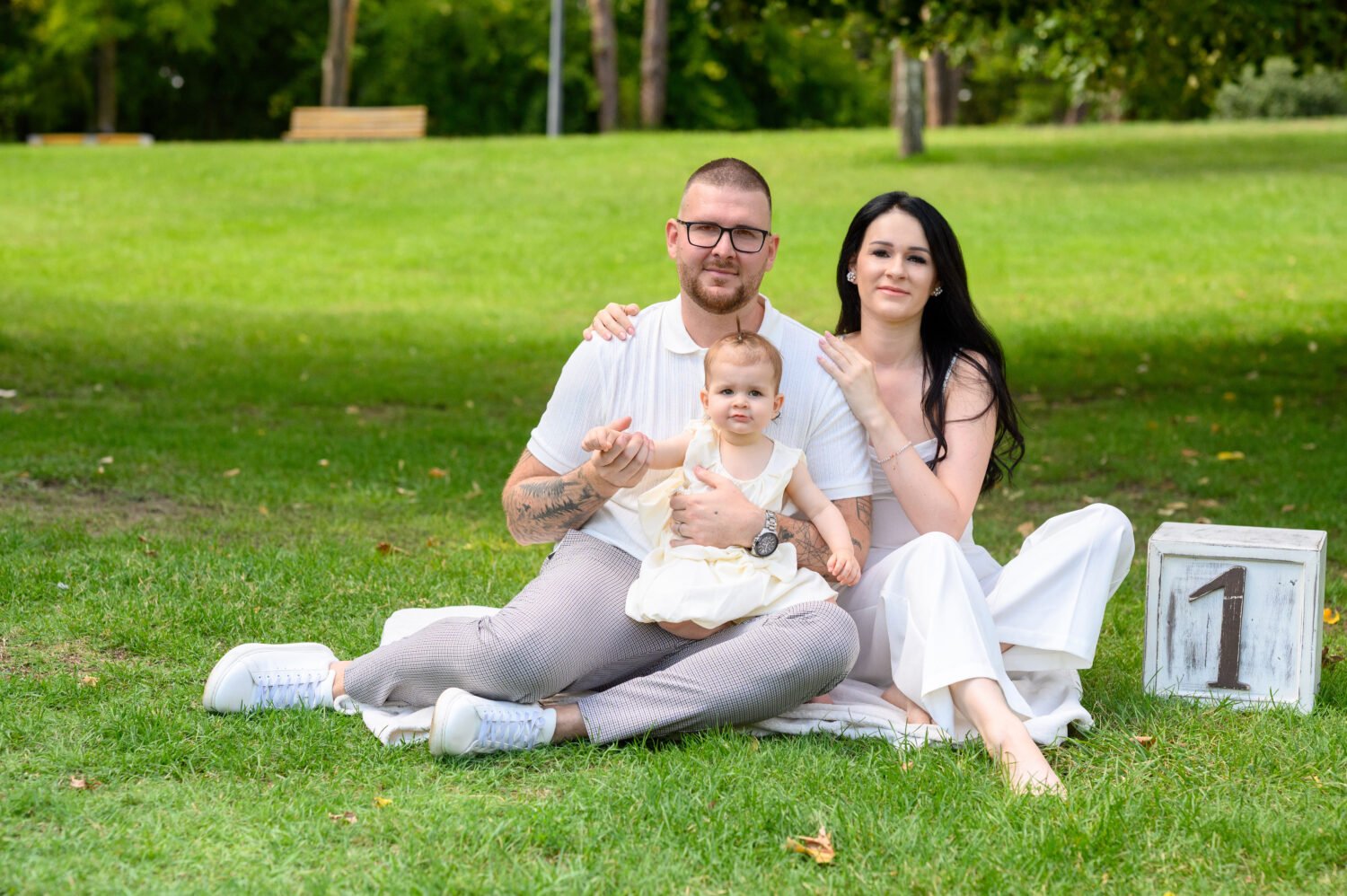 Anya, apa és egyéves kislányuk a park füvén ülve, mellettük fehér fa kocka egyes számmal, szabadtéri fotózás.