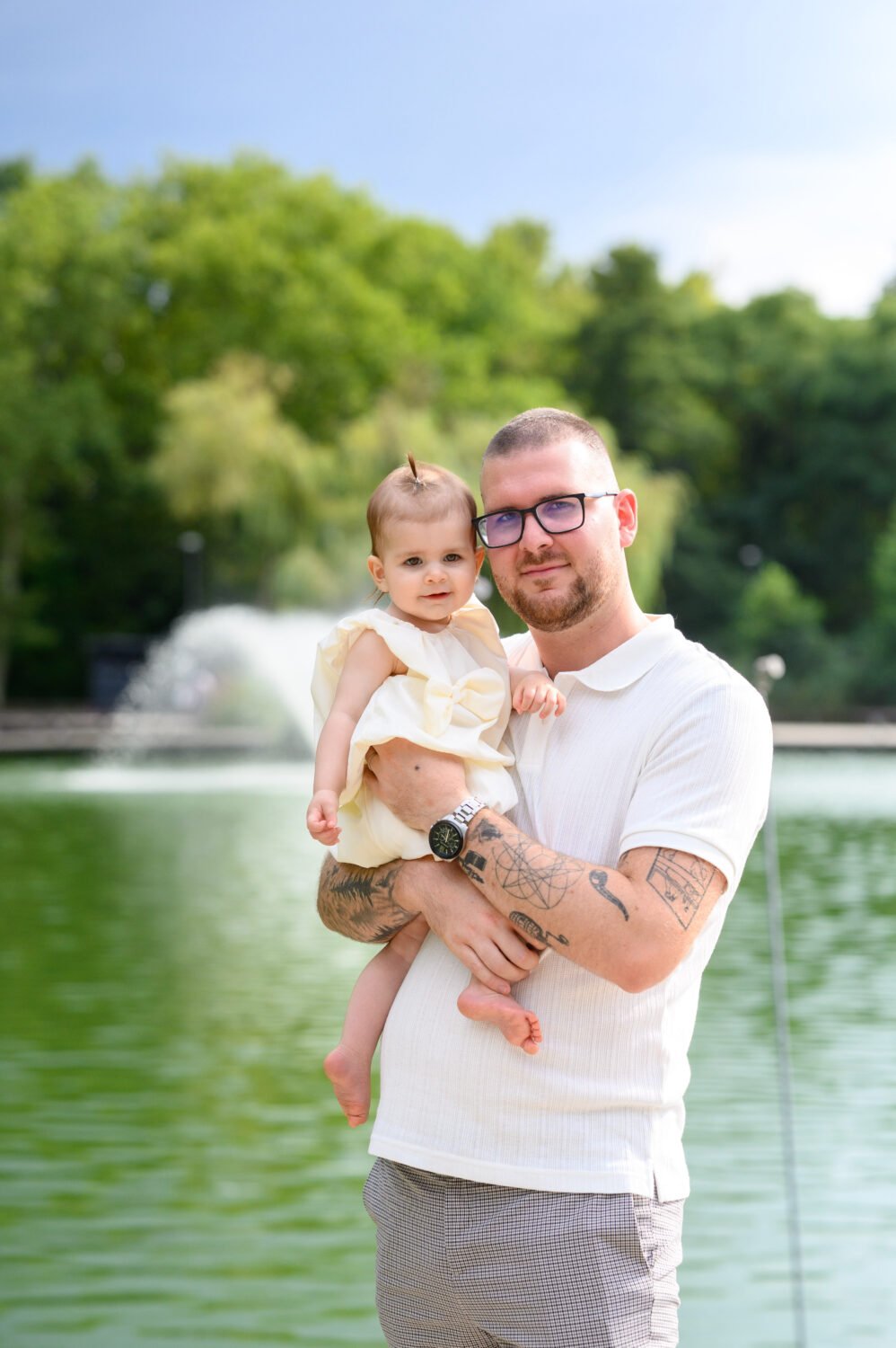 Szemüveges édesapa fehér pólóban tartja egyéves kislányát egy tó partján, háttérben szökőkút és park.