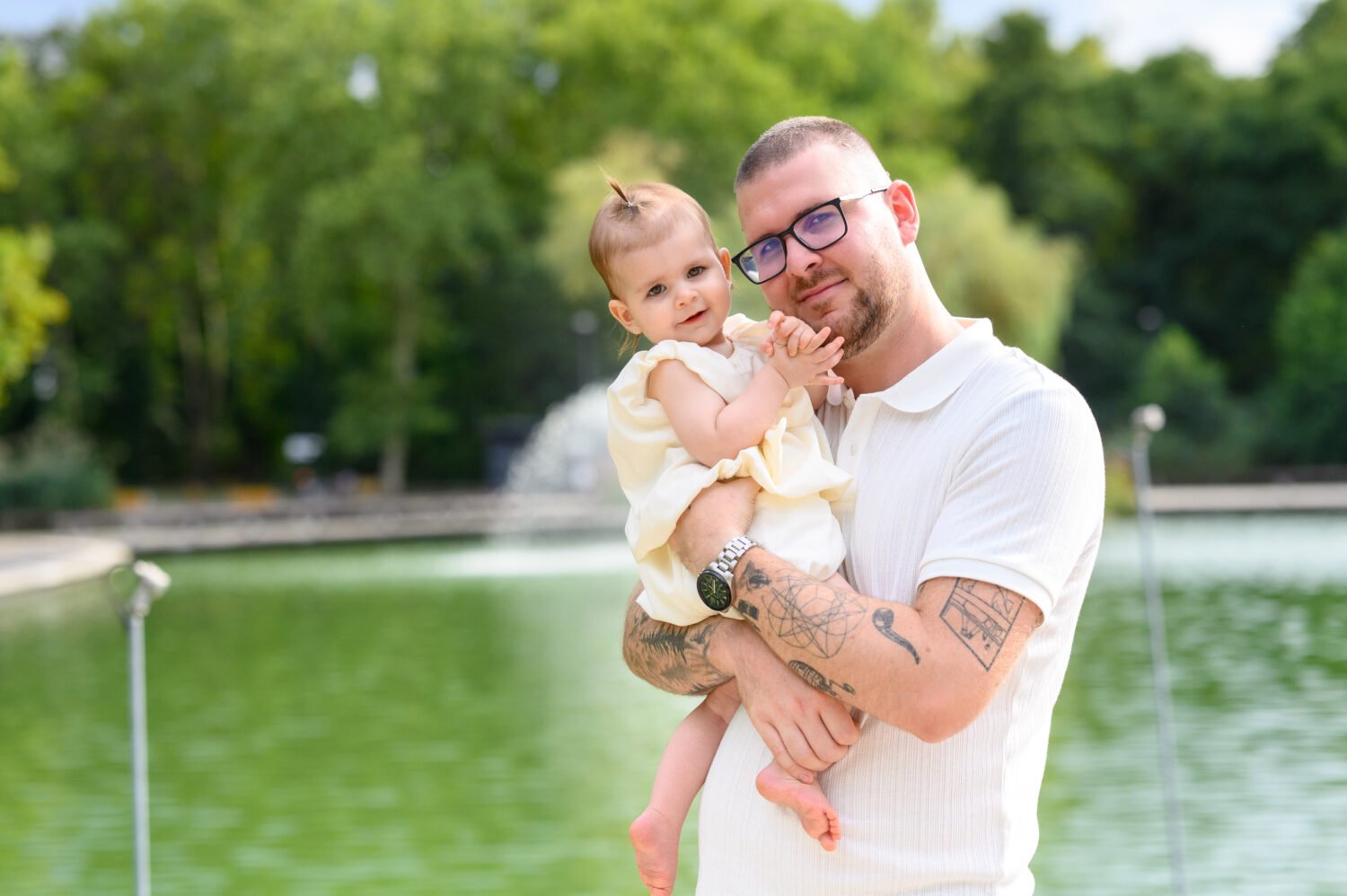 Édesapa a karjában tartja mosolygós egyéves kislányát egy tó partján, háttérben szökőkút és zöld park.