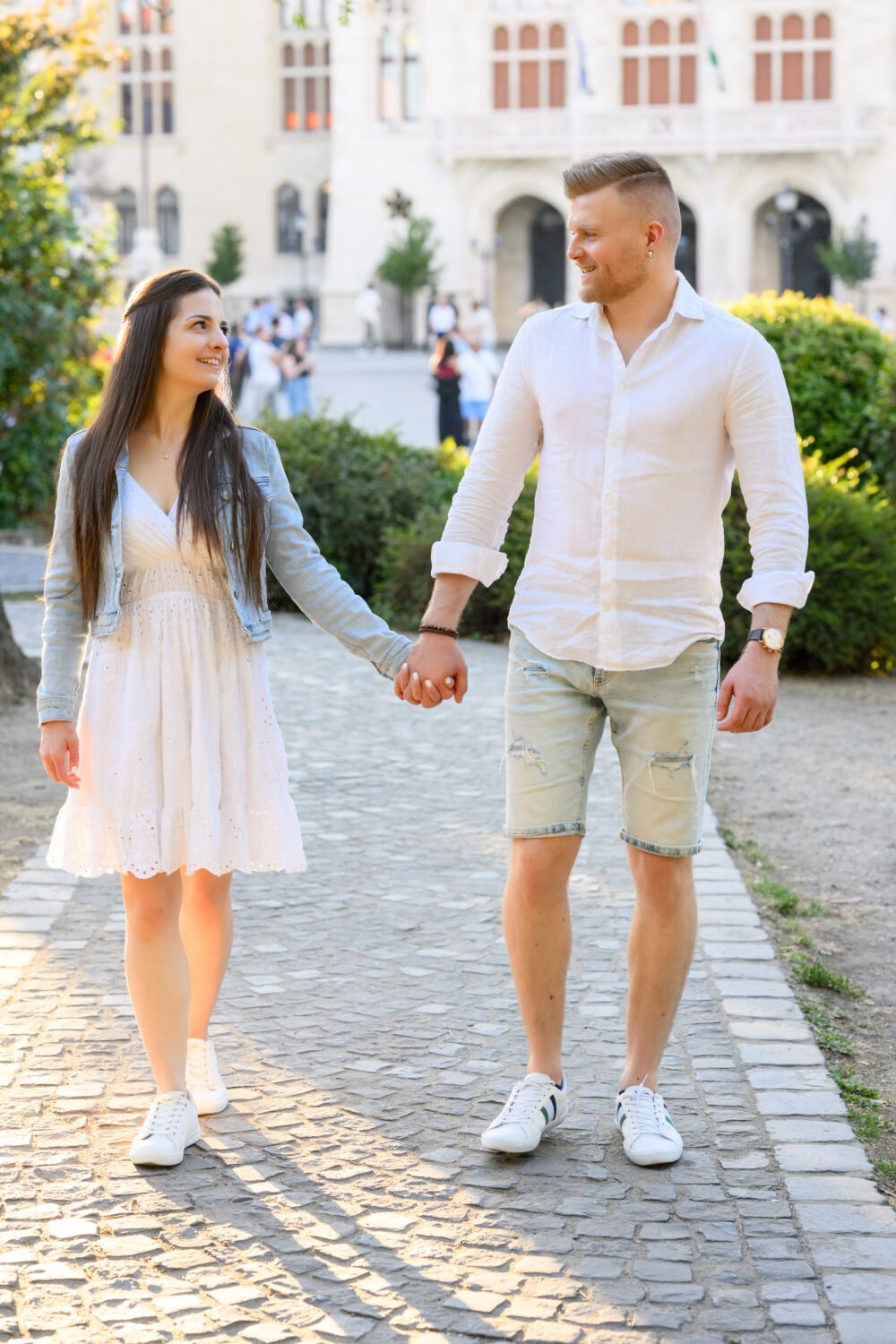 Jegyespár kéz a kézben sétál egy budapesti kastélykert macskaköves útján az aranyóra fényében, egymásra mosolyogva – nyári jegyes fotózás.