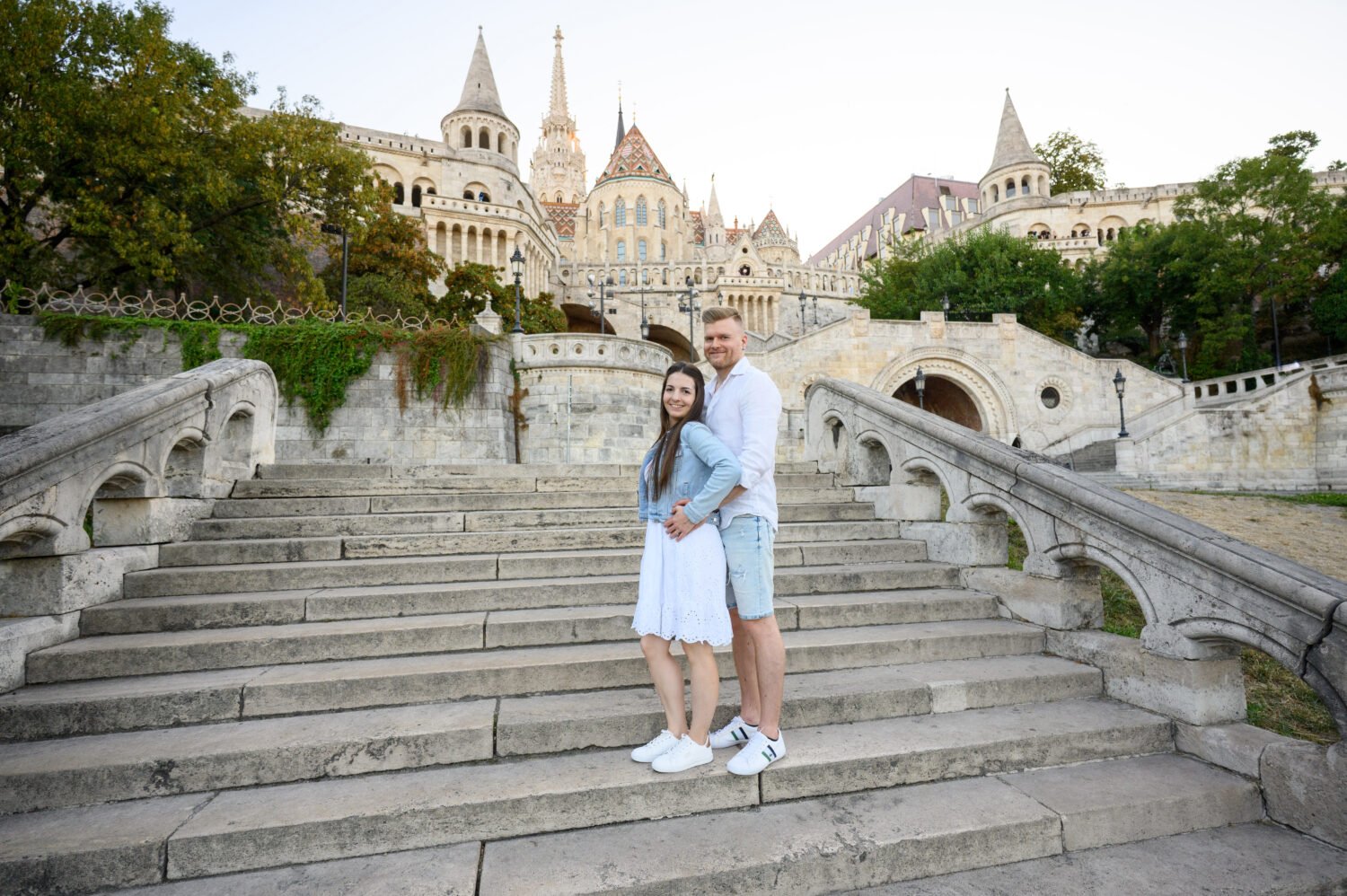 Jegyespár összebújva ül a Halászbástya széles kőlépcsőjén, háttérben a fehér tornyok és a Mátyás-templom – budapesti jegyes fotózás.