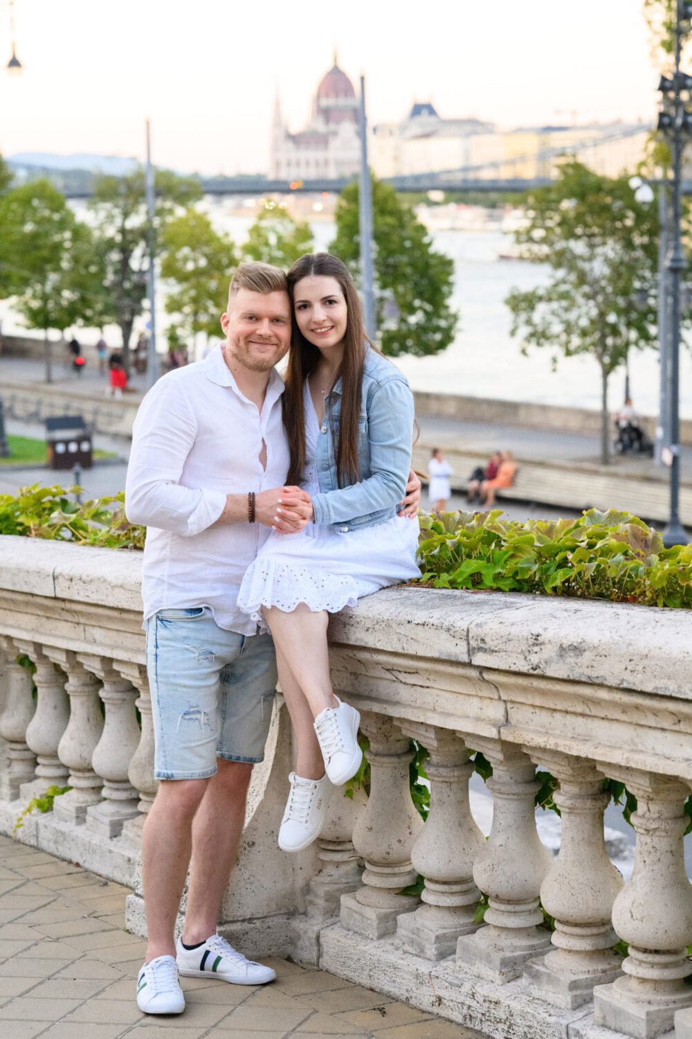 Jegyespár egy kő korláton összebújva, háttérben a Duna, a Parlament és a Lánchíd naplementében – budapesti jegyes fotózás.